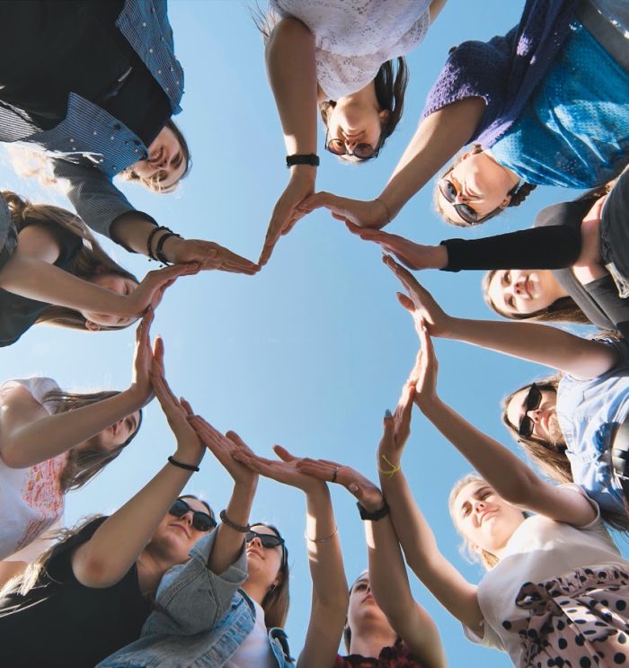Gruppe von Menschen steht im Kreis und legt die Hände zusammen als Symbol für Teamarbeit, Zusammenarbeit und Gemeinschaft unter blauem Himmel.